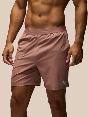 A close-up of a man's lower torso wearing dusty rose athletic shorts with a small white wing logo on the lower left leg, set against a solid beige background.