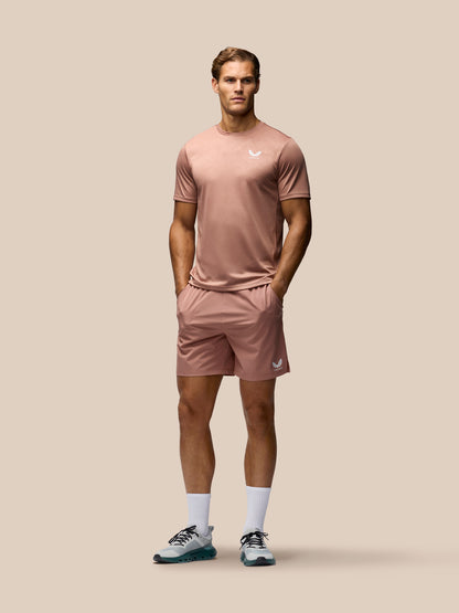 A male model stands against a solid beige background wearing a matching dusty rose athletic set consisting of a short-sleeve t-shirt and shorts, paired with white socks and running shoes.