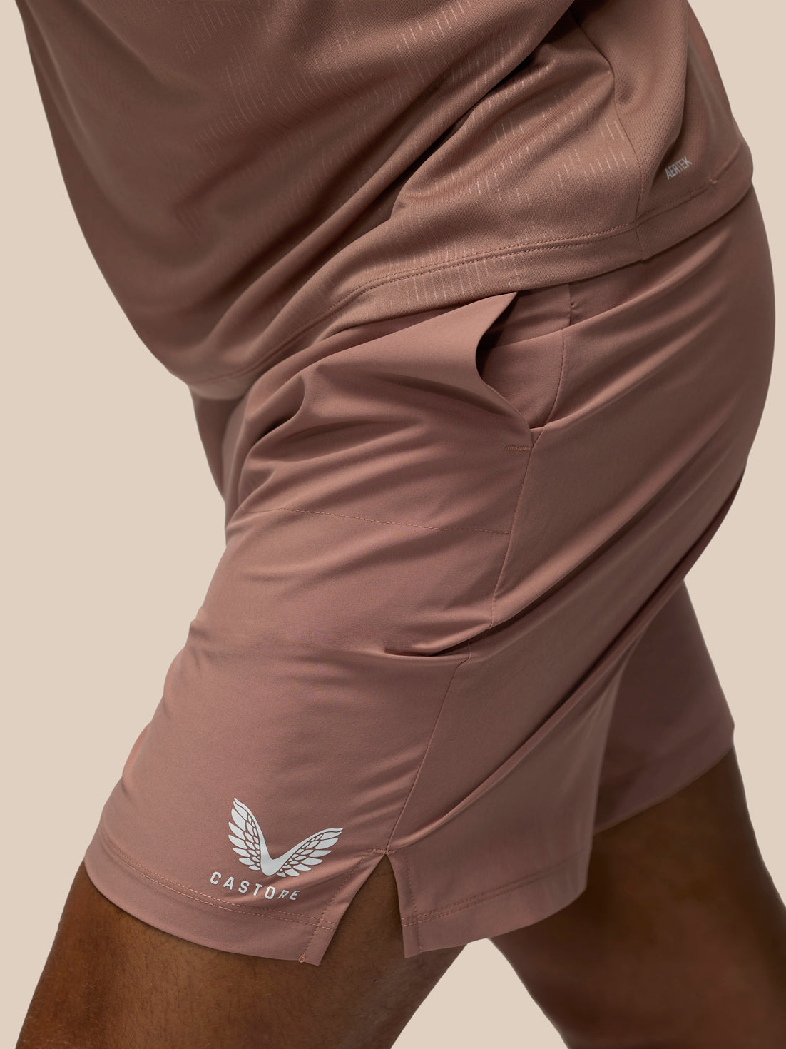 Close-up of a person wearing dusty rose athletic shorts with a white winged logo and the brand name CASTORE on the lower left leg, paired with a matching top against a plain beige background.