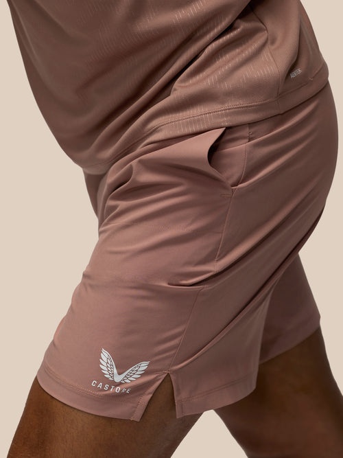 Close-up of a person wearing dusty rose athletic shorts with a white winged logo and the brand name CASTORE on the lower left leg, paired with a matching top against a plain beige background.