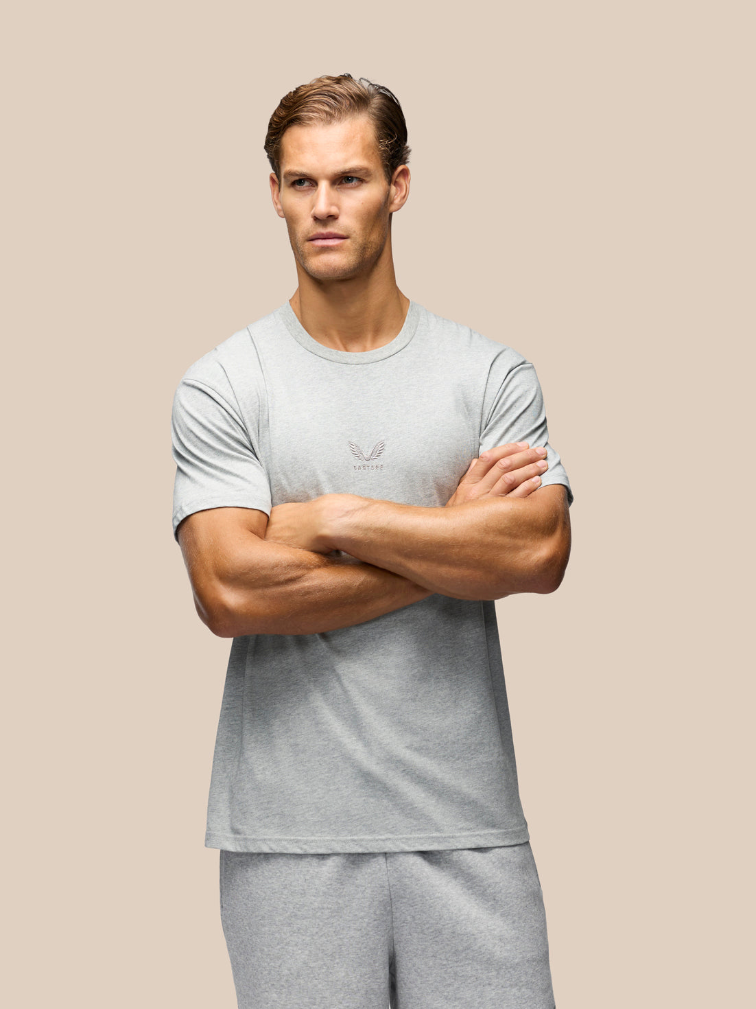 A muscular man wearing a light gray short-sleeve athletic t-shirt and matching gray sweatpants, standing with arms crossed against a solid beige background.