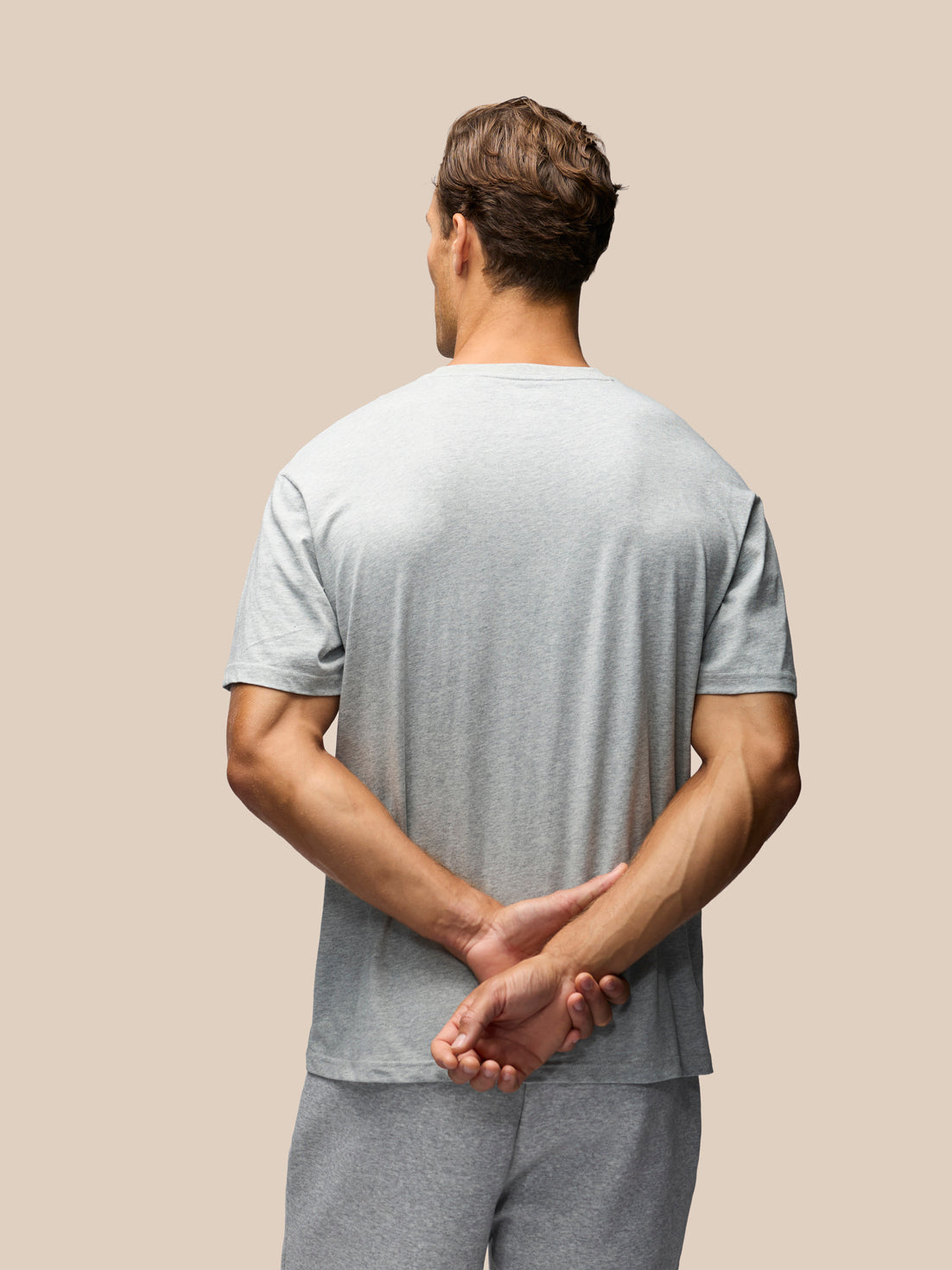 A man viewed from behind wearing a light gray short-sleeve t-shirt and matching gray sweatpants, standing against a solid beige background with his hands clasped behind his back.