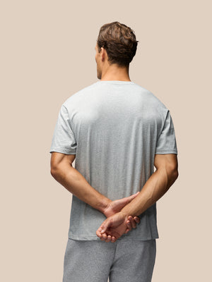 A man viewed from behind wearing a light gray short-sleeve t-shirt and matching gray sweatpants, standing against a solid beige background with his hands clasped behind his back.