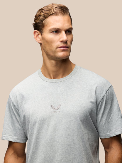 A man wearing a light gray crewneck t-shirt with a small embroidered winged logo on the center chest, set against a solid beige background.