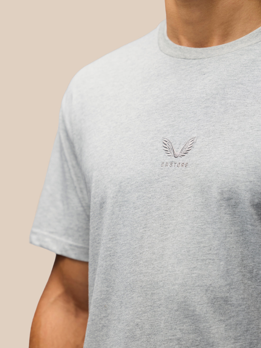 A close-up of a person wearing a light heather gray crewneck t-shirt with a small winged logo and 'CASTORE' text on the left chest, set against a solid beige background.