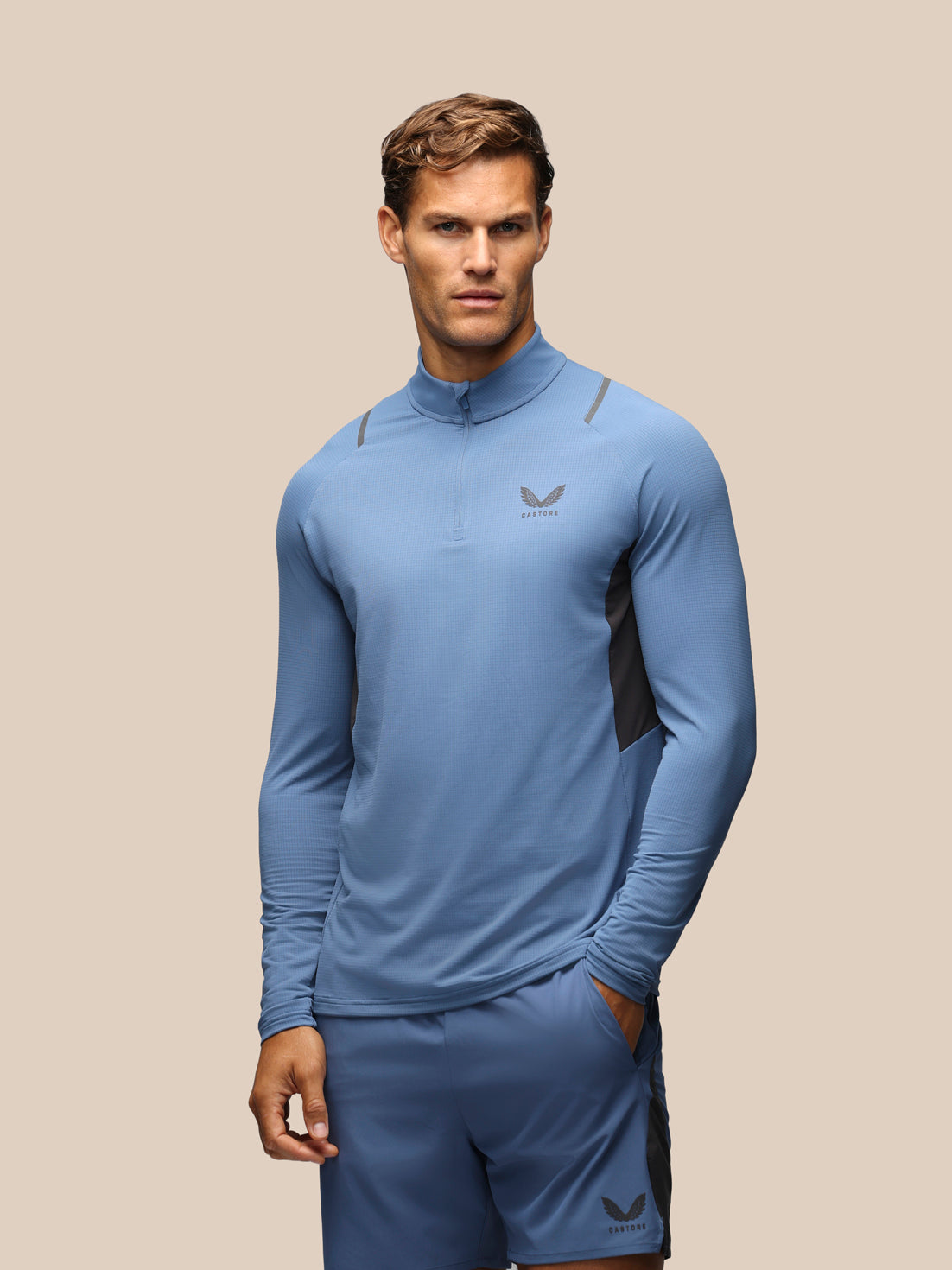 A male model wearing a light blue athletic quarter-zip pullover and matching shorts with dark gray side panels and logos, set against a solid beige background.
