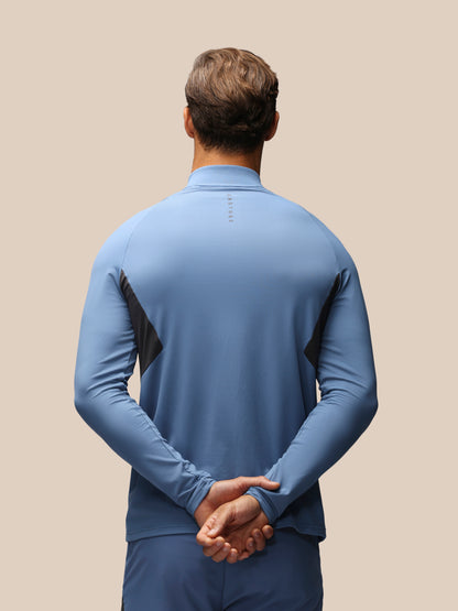 A man viewed from behind wearing a light blue long-sleeve athletic quarter-zip top with black side panels and matching blue athletic pants, set against a solid beige background.