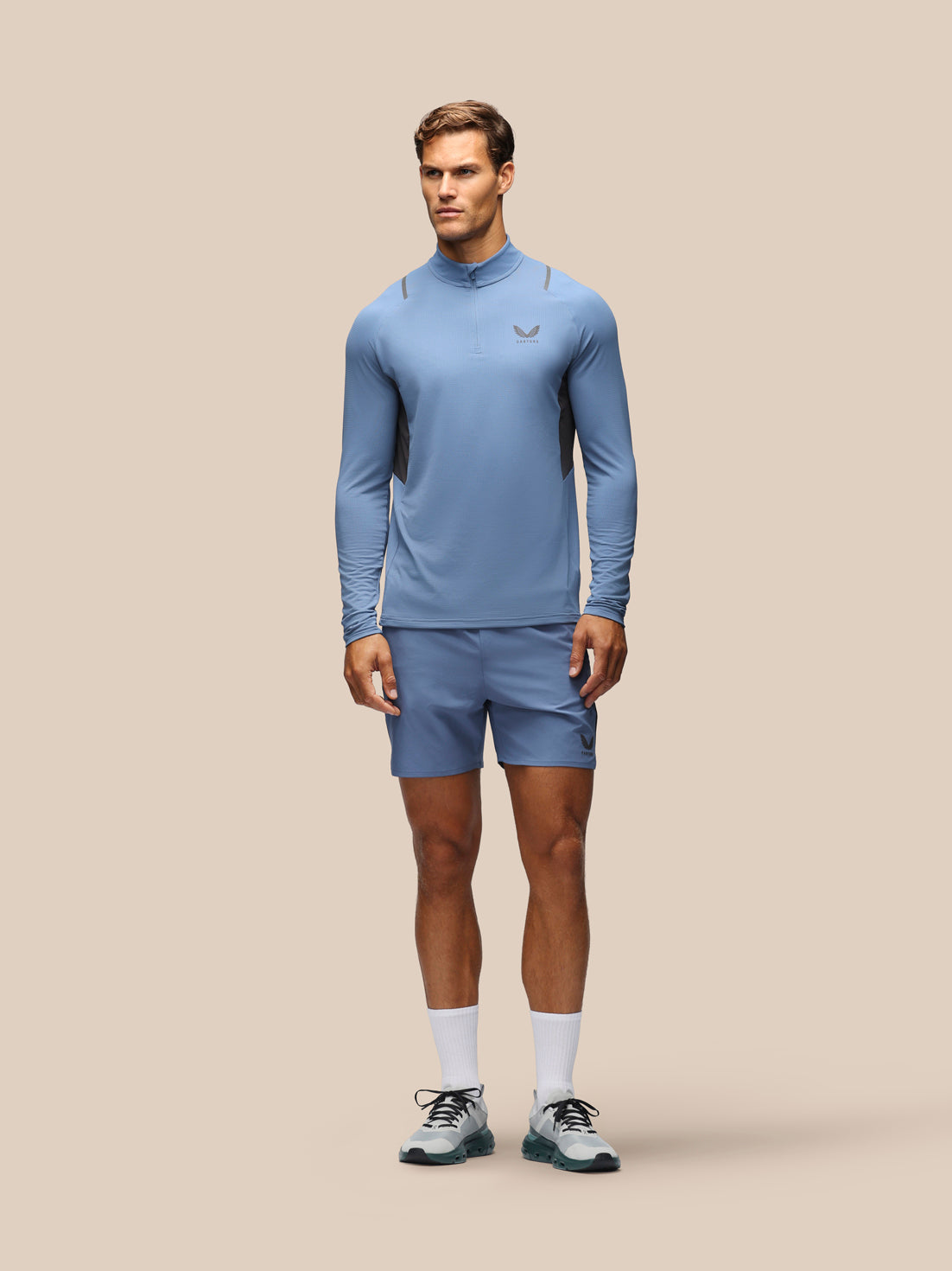 A male model stands against a solid beige background wearing a light blue athletic long-sleeve mock-neck top and matching shorts, paired with white socks and gray running shoes.