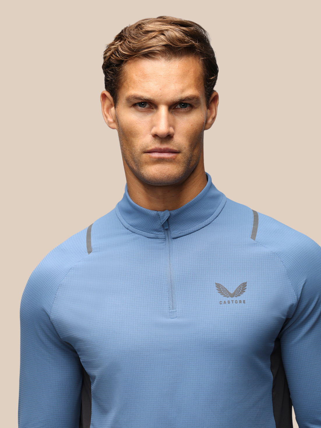 A man wearing a light blue long-sleeve athletic quarter-zip top with a small gray wing logo and the word 'CASTORE' on the left chest, photographed against a plain beige studio background.