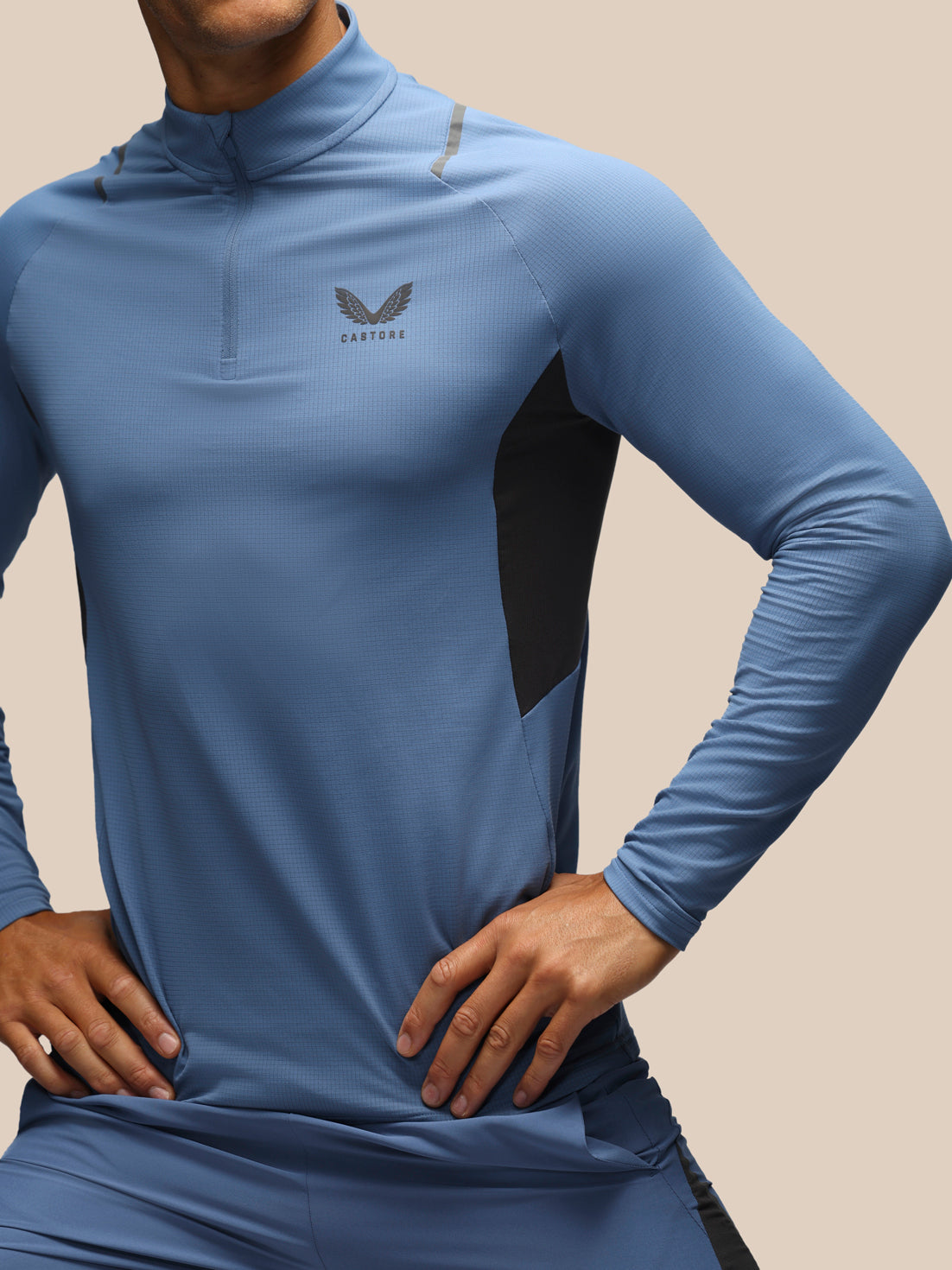 A close-up of a man wearing a blue long-sleeve athletic quarter-zip top with black side panels and a gray wing logo on the chest, set against a solid beige background.