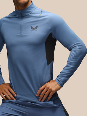 A close-up of a man wearing a blue long-sleeve athletic quarter-zip top with black side panels and a gray wing logo on the chest, set against a solid beige background.
