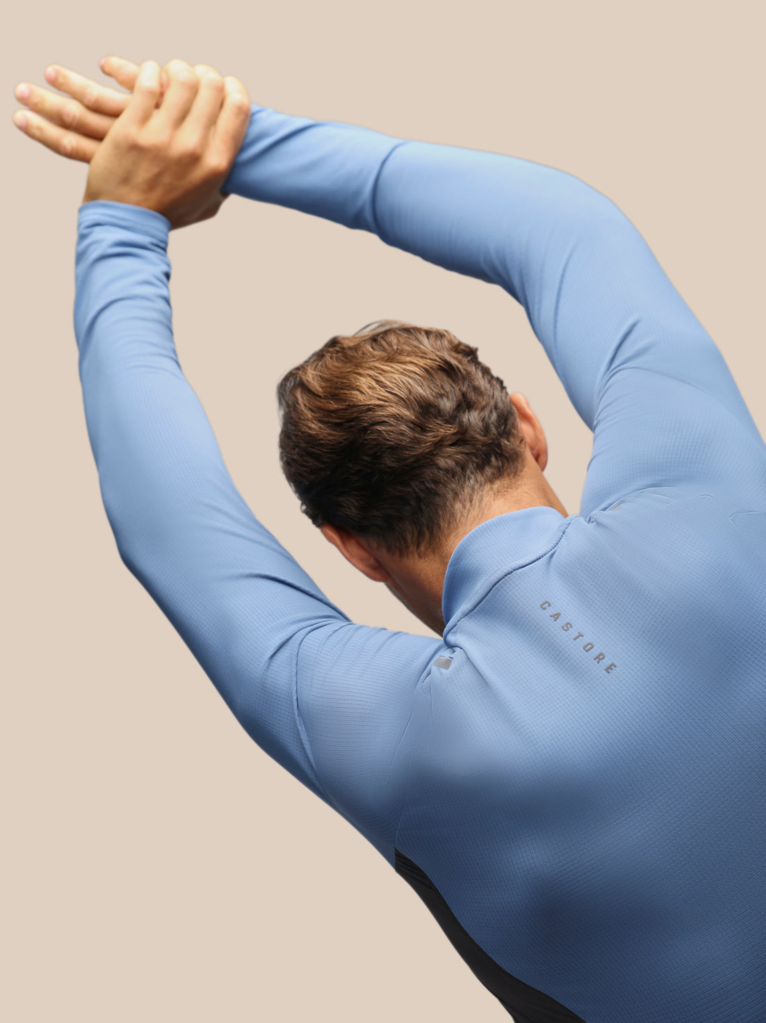 A man wearing a light blue long-sleeve performance top, shown from behind as he stretches his arms overhead. The brand name 'CASTORE' is subtly printed on the upper back.