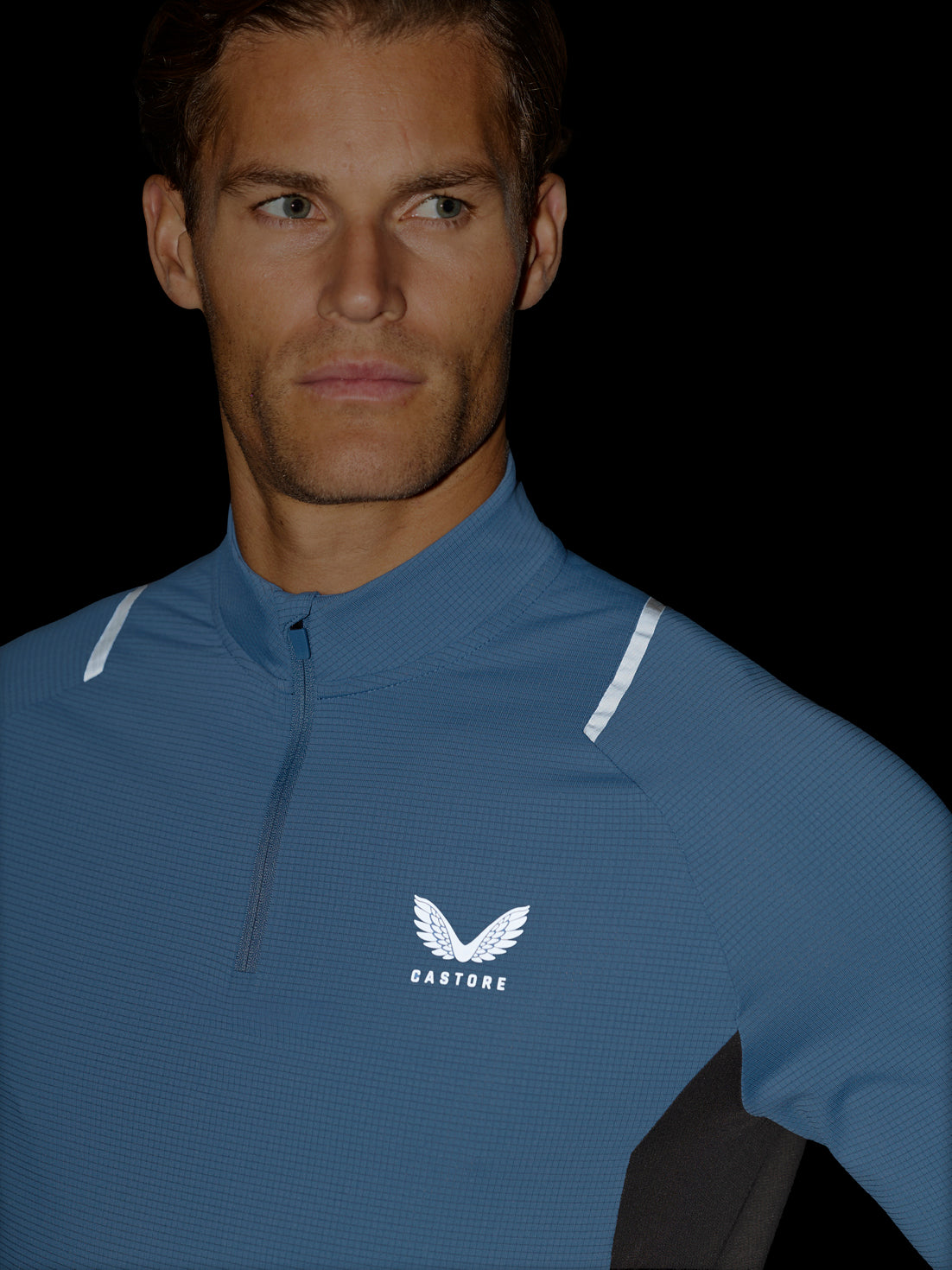 A close-up of a man wearing a blue athletic quarter-zip pullover with a white wing logo and the brand name CASTORE on the chest, set against a solid black background.