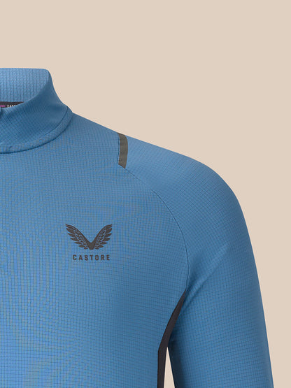 Close-up of a light blue long-sleeve athletic top with a dark gray wing logo and the word CASTORE on the chest, set against a solid beige background.