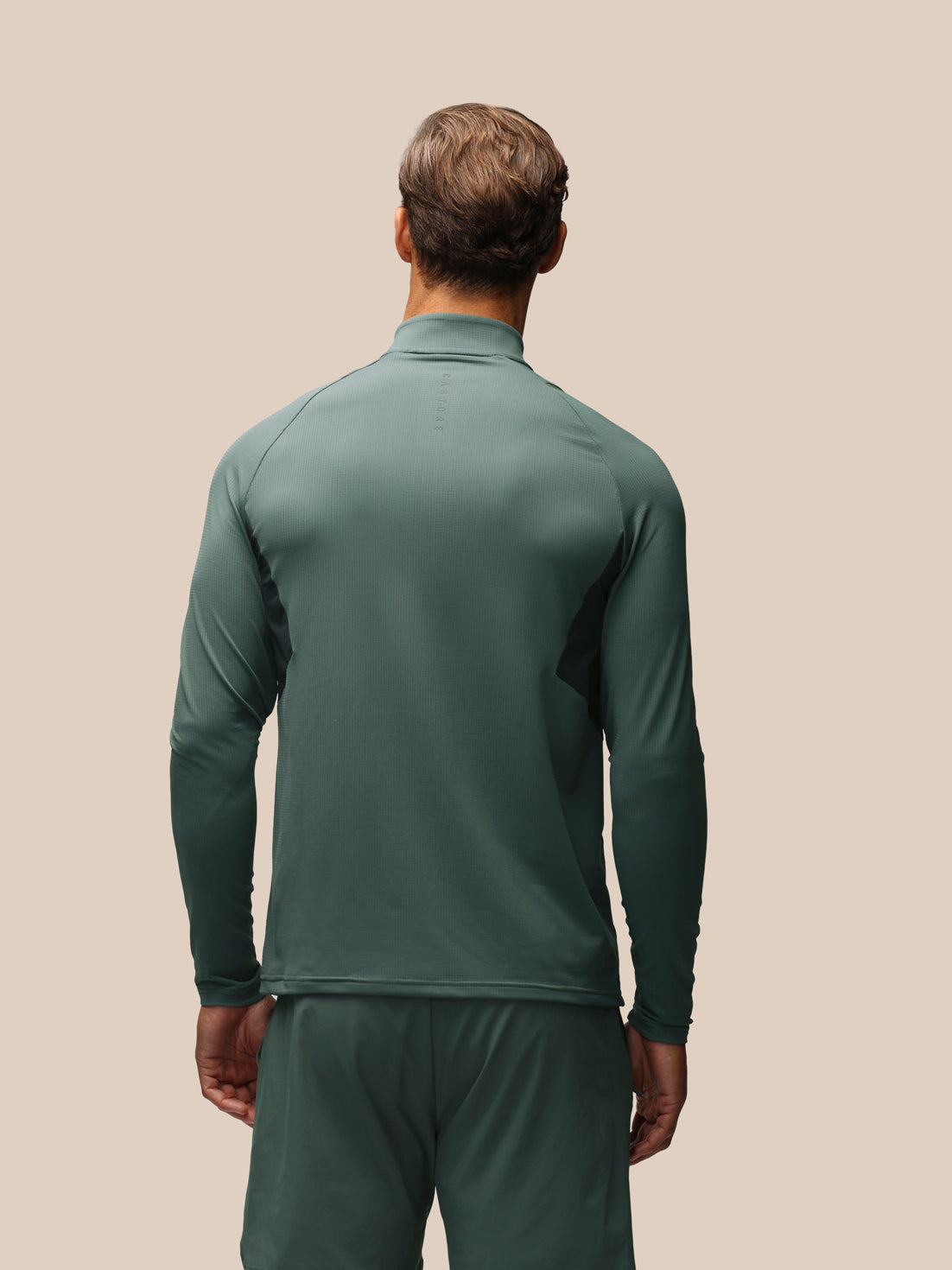 A man viewed from behind wearing a dark green long-sleeve athletic quarter-zip pullover and matching green pants against a solid beige background.