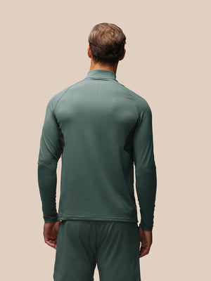 A man viewed from behind wearing a dark green long-sleeve athletic quarter-zip pullover and matching green pants against a solid beige background.
