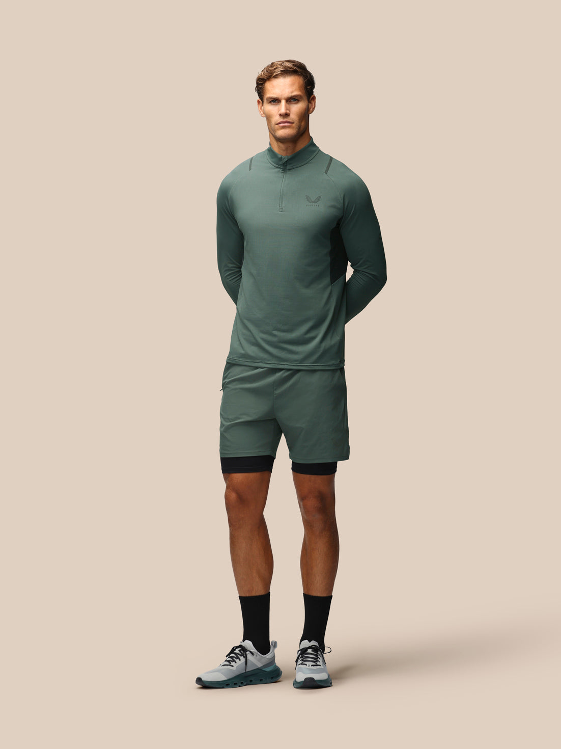 A male model stands against a solid beige background wearing a matching athletic set consisting of a long-sleeve quarter-zip pullover and matching shorts in a muted sage green color, styled with black compression sleeves and gray running shoes.