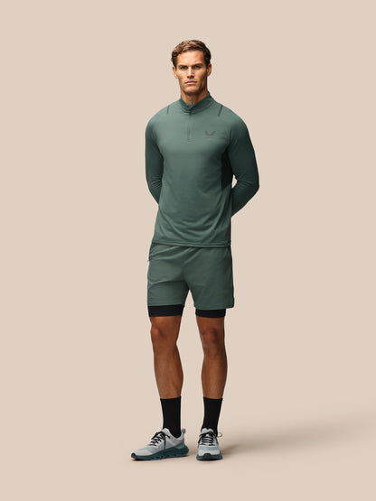 A male model stands against a solid beige background wearing a matching athletic set consisting of a long-sleeve quarter-zip pullover and matching shorts in a muted sage green color, styled with black compression sleeves and gray running shoes.