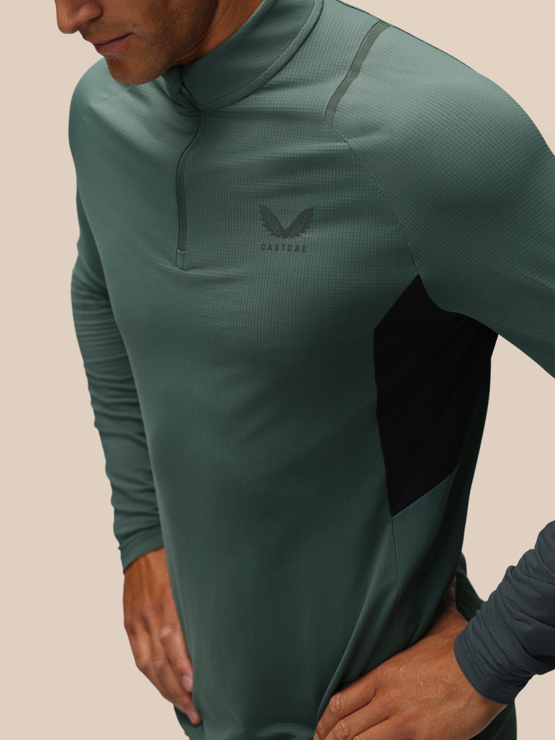 A close-up of a man wearing a dark green ribbed quarter-zip athletic pullover with a black side panel and a subtle wing logo on the chest.