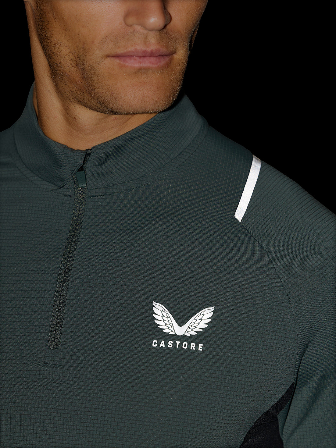 Close-up of a man wearing a dark gray Castore quarter-zip pullover jacket with a white wing logo and the brand name 'CASTORE' on the chest, set against a solid black background.