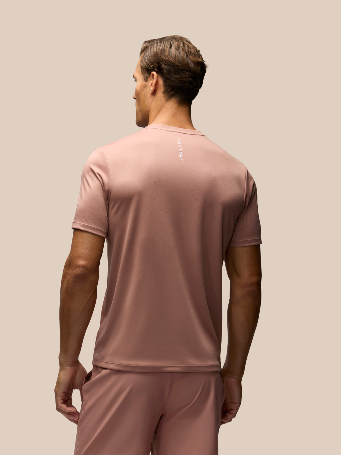 A man viewed from behind wearing a dusty rose short-sleeve athletic t-shirt and matching shorts against a solid beige background.
