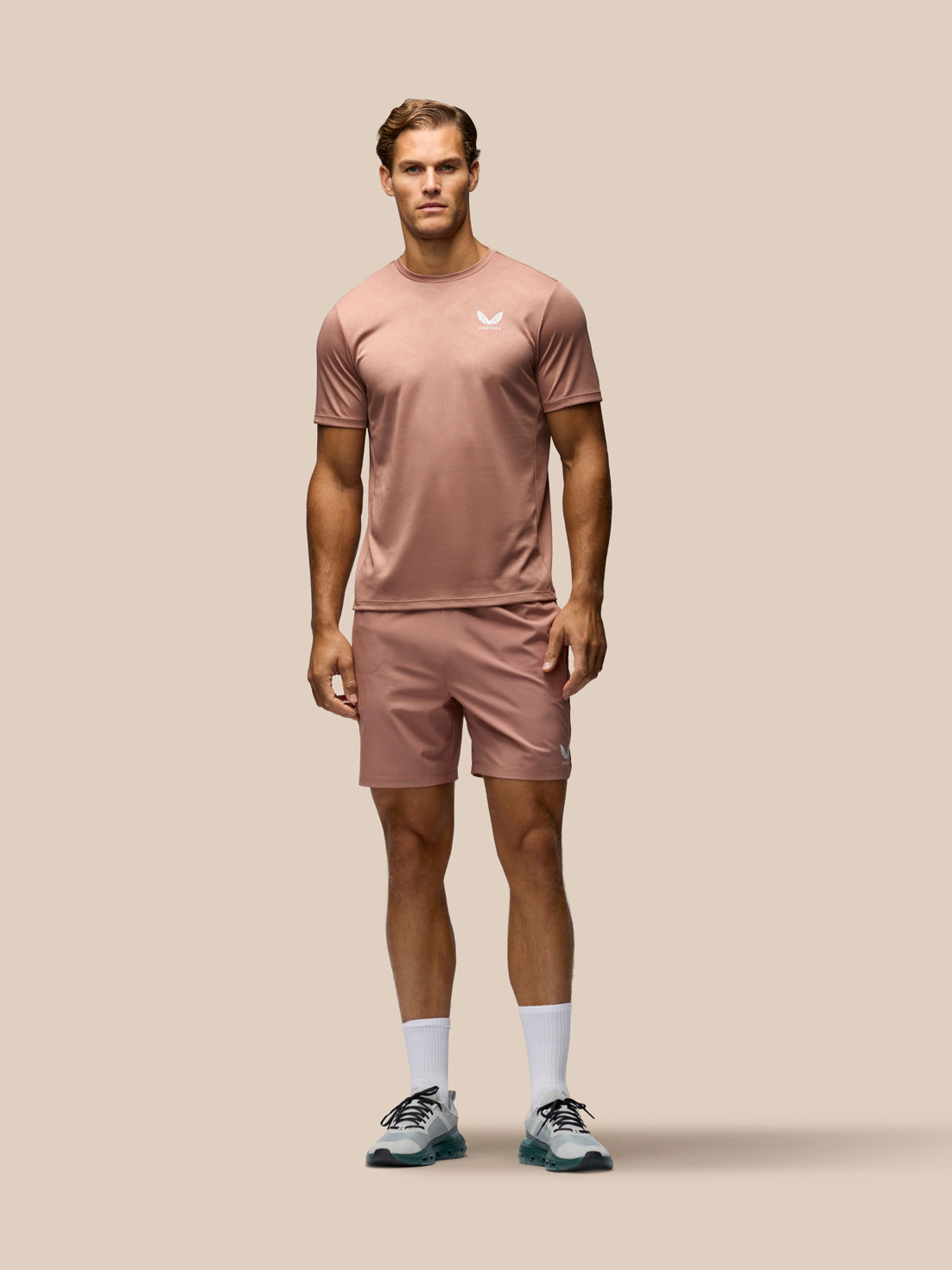 A man modeling a matching dusty rose athletic set consisting of a short-sleeve crewneck t-shirt and matching shorts, with white socks and athletic shoes, against a solid beige background.