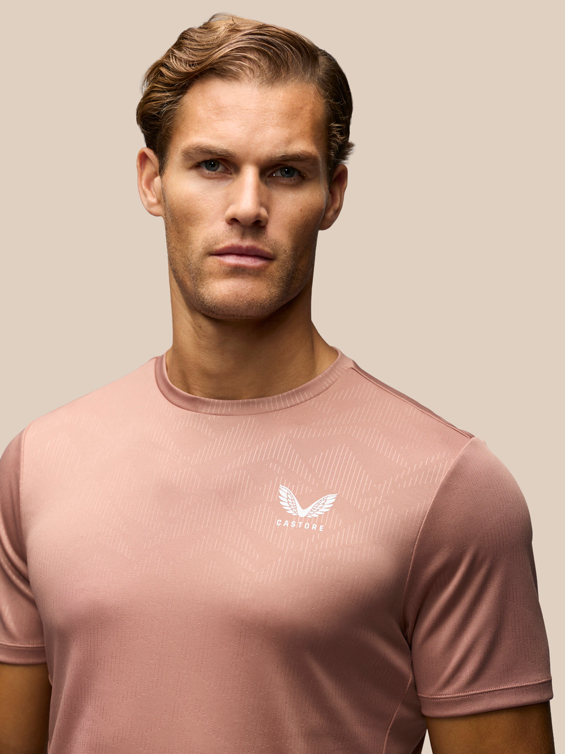 A man wearing a dusty rose pink athletic t-shirt with a white winged logo and the brand name 'CASTORE' on the left chest, set against a solid beige background.