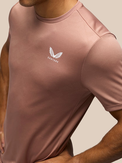 A close-up of a man wearing a dusty rose short-sleeve athletic t-shirt with a white winged logo and the brand name 'CASTORE' printed on the left chest, set against a solid beige background.