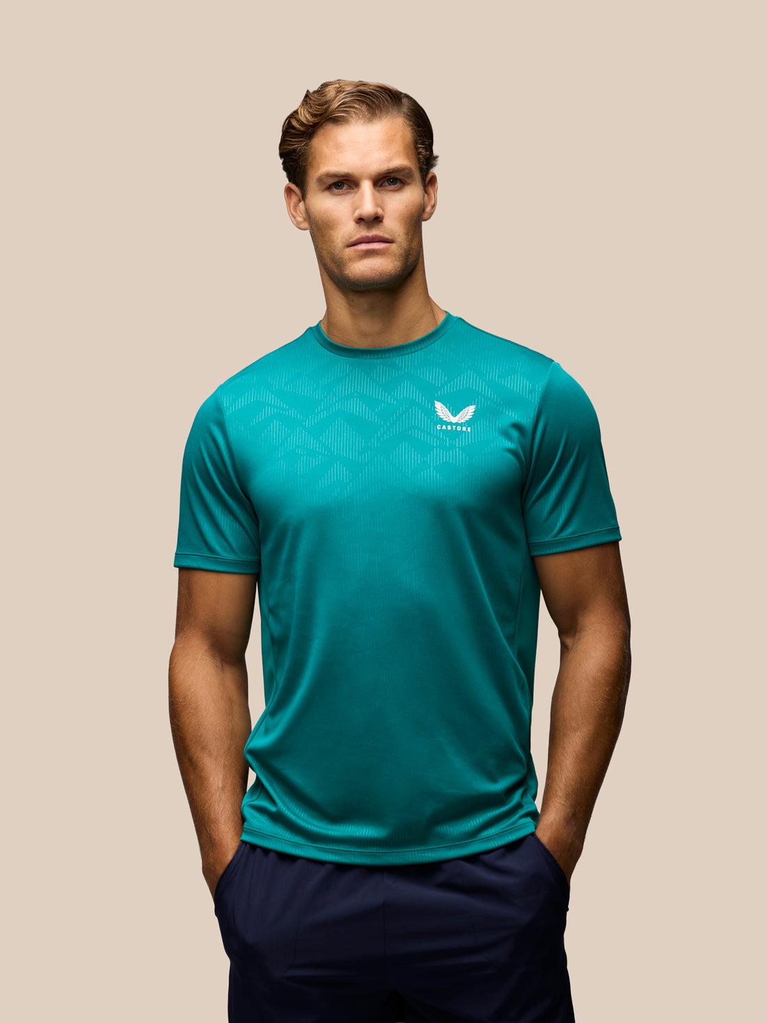 A man wearing a teal short-sleeve athletic t-shirt with a white wing logo on the left chest, paired with dark navy pants, set against a solid beige background.