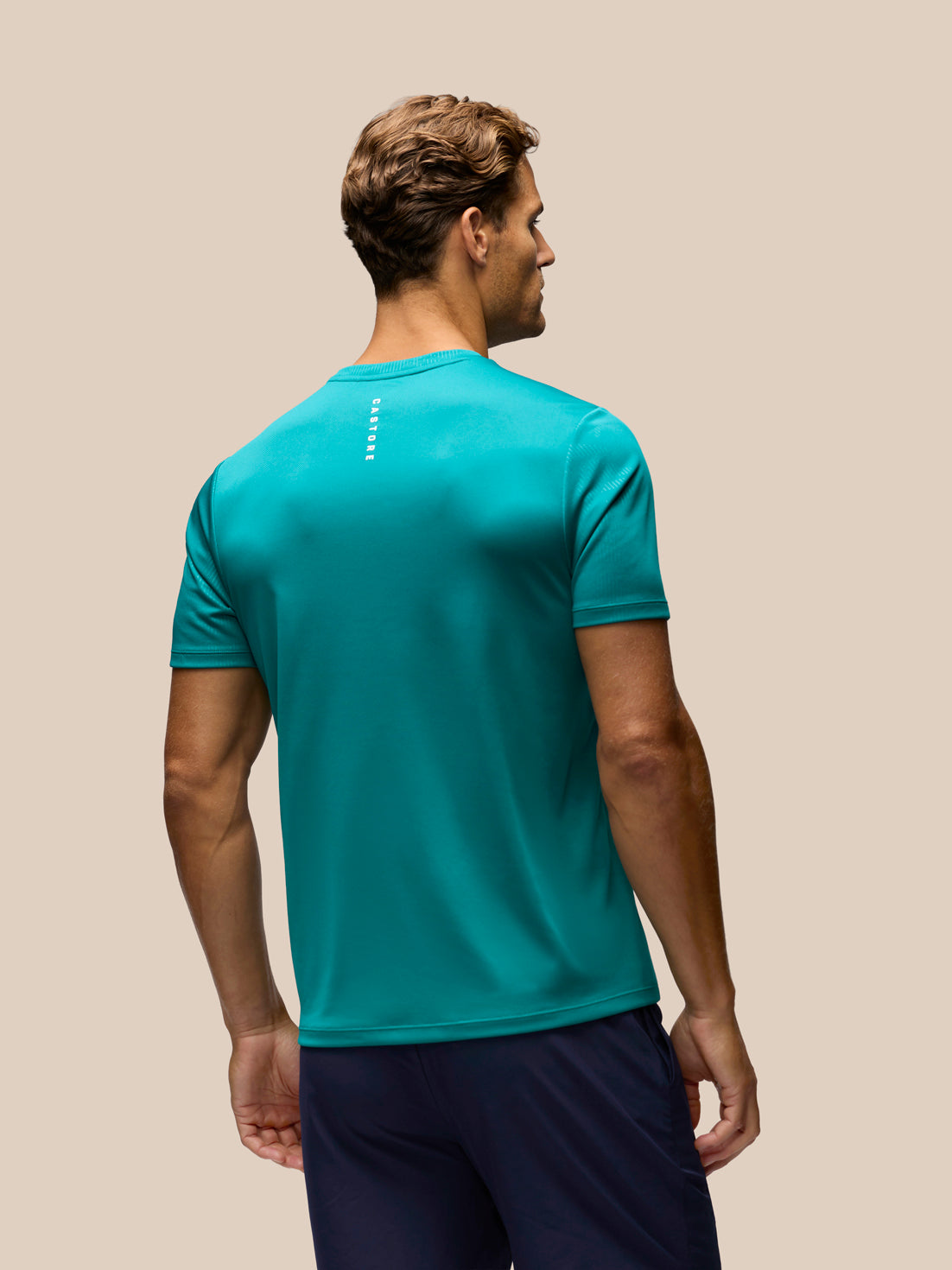 A man wearing a teal athletic t-shirt and navy shorts, shown from the back against a solid beige background.