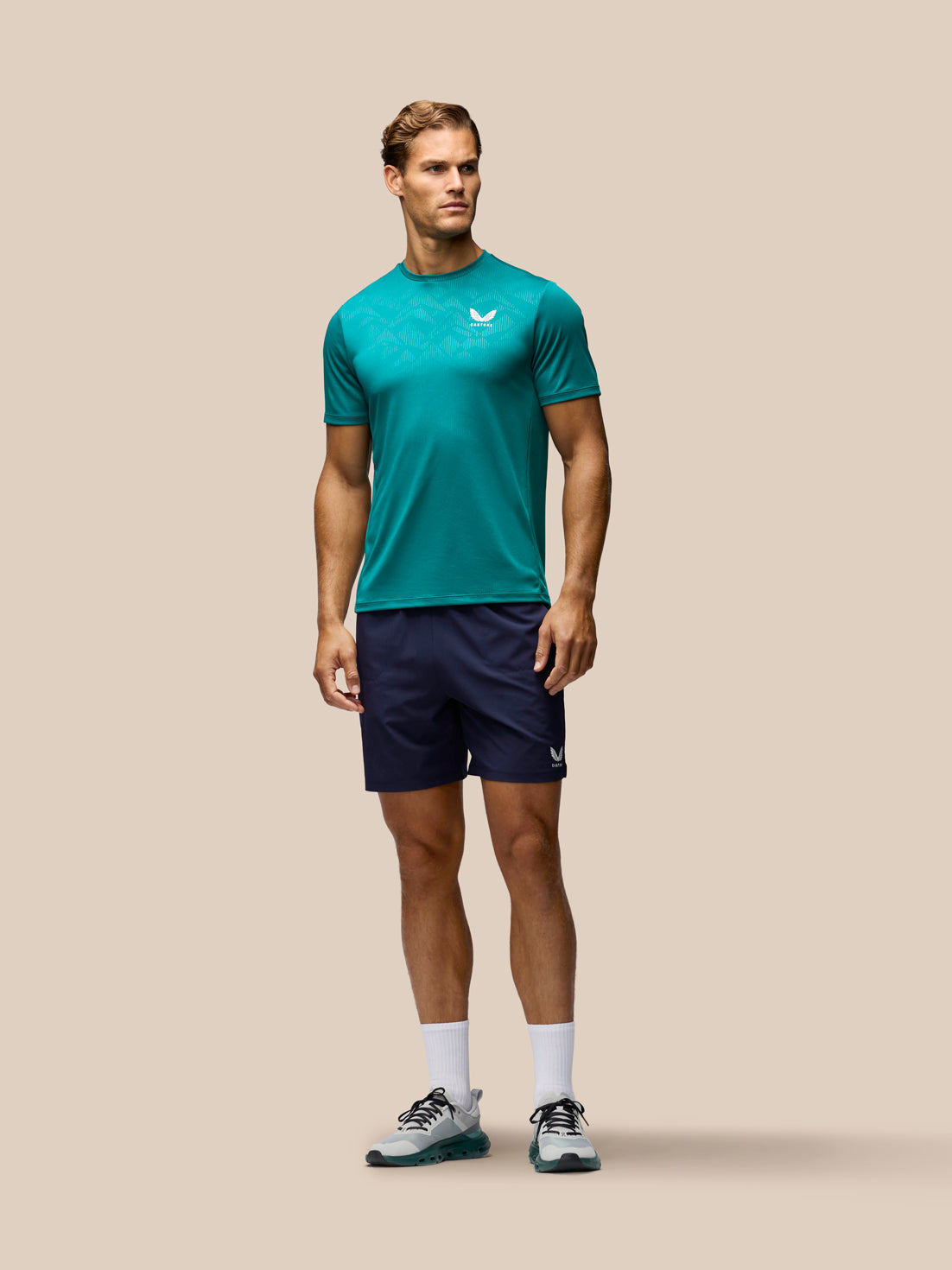 A male model stands against a solid beige background wearing a teal athletic t-shirt with a white logo, navy blue athletic shorts, white socks, and gray running shoes.
