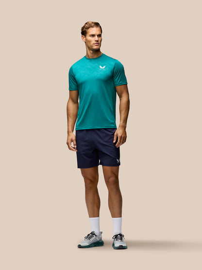 A male model stands against a solid beige background wearing a teal athletic t-shirt with a white logo, navy blue athletic shorts, white socks, and gray running shoes.