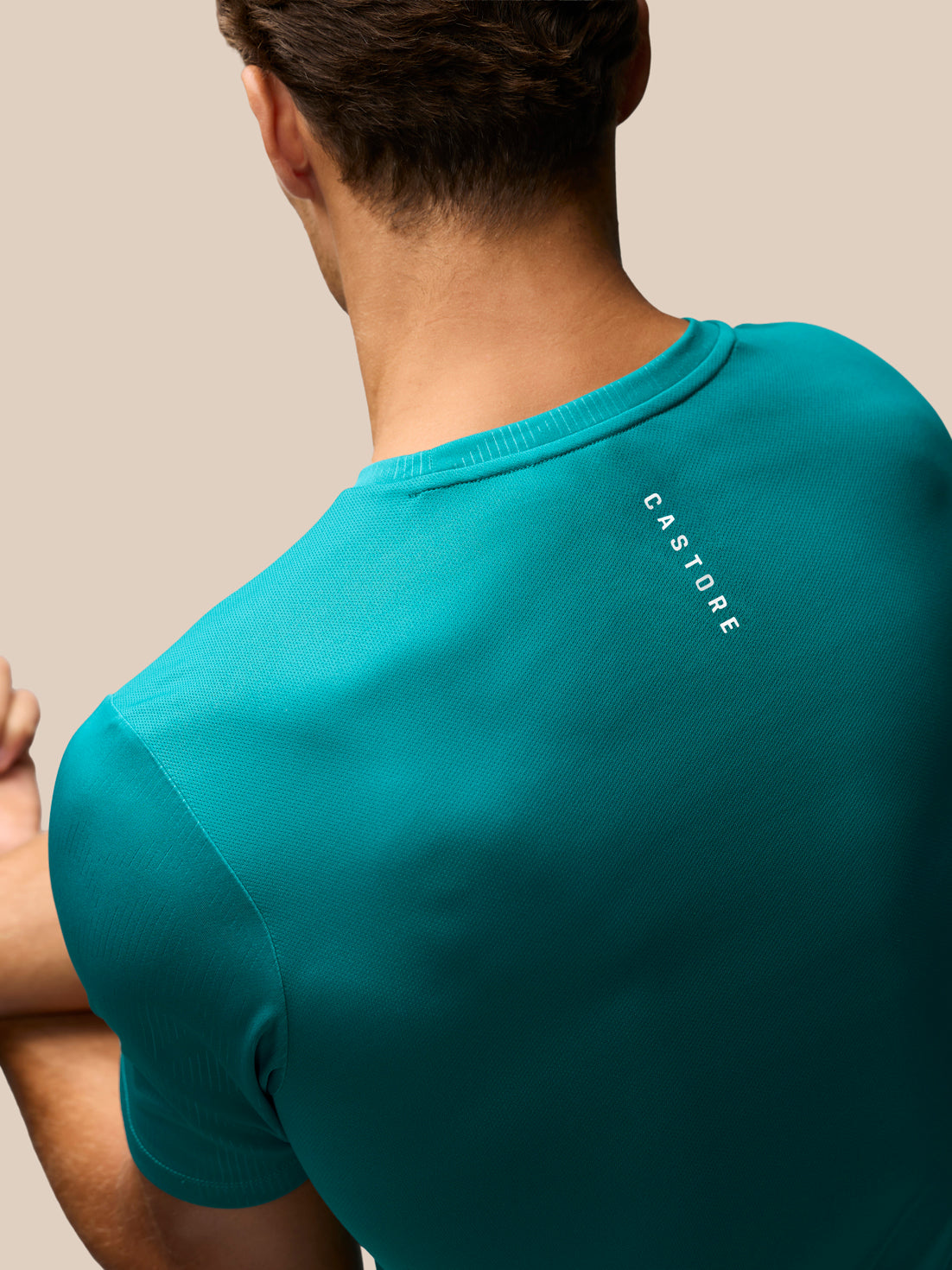 A man wearing a teal short-sleeve t-shirt, shown from the back over his shoulder. The shirt features the brand name 'CASTORE' printed vertically in white on the upper right shoulder.
