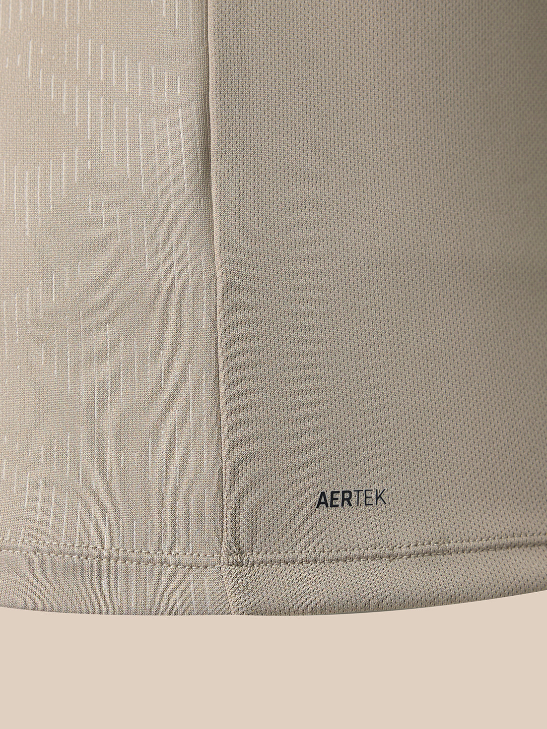 Close-up of a light beige athletic fabric with a subtle vertical textured pattern and a small black AERTEK logo near the bottom hem.