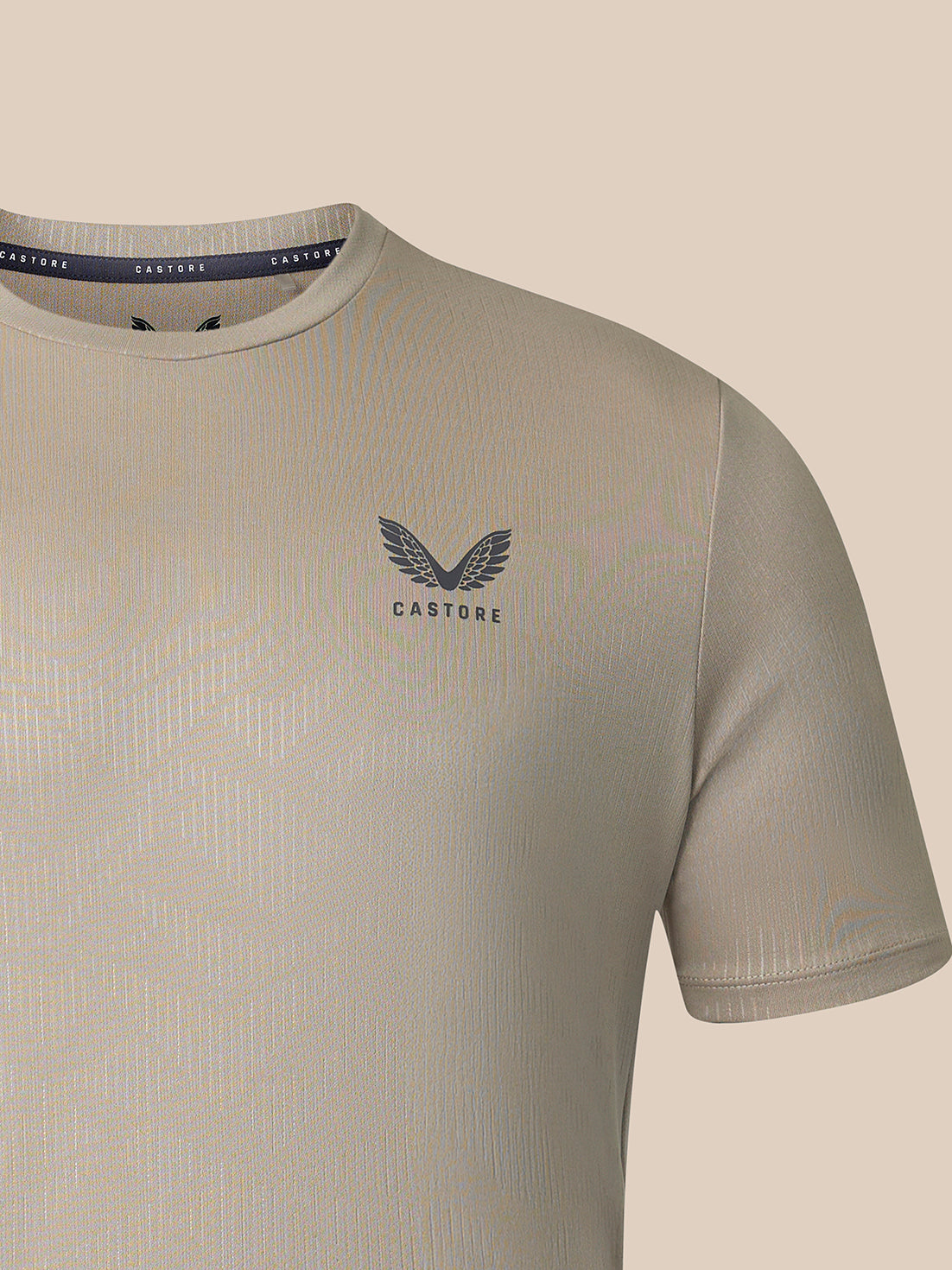 Close-up of a Castore short-sleeve t-shirt in a light beige-gray color, featuring a dark gray winged logo and the brand name 'CASTORE' printed on the chest, set against a solid beige background.