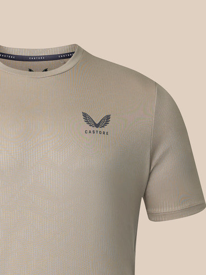 Close-up of a Castore short-sleeve t-shirt in a light beige-gray color, featuring a dark gray winged logo and the brand name 'CASTORE' printed on the chest, set against a solid beige background.