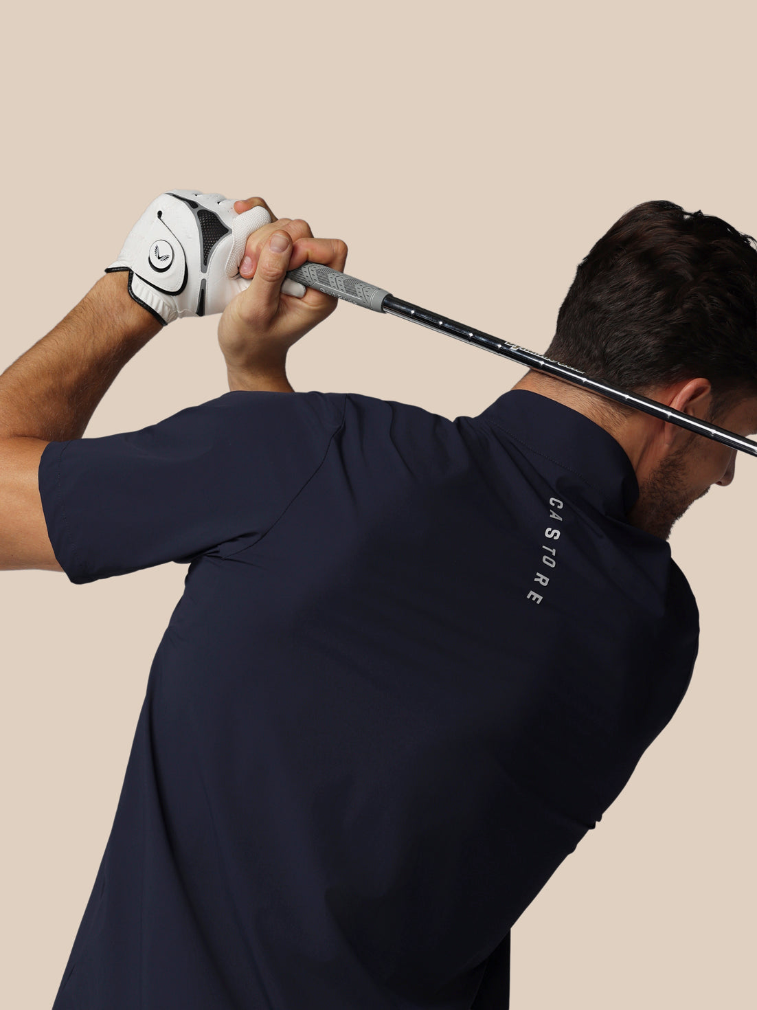 Man in navy blue shirt with 'Castore' branding holding a golf club against a beige background