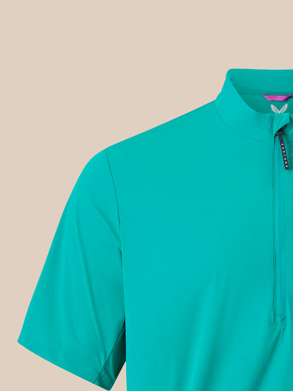 A close-up of a short-sleeve athletic half-zip pullover in a bright turquoise color, featuring a mock neck collar and a black zipper pull with white branding, set against a solid beige background.