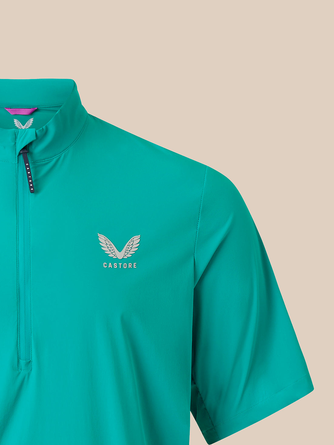 A close-up of a teal short-sleeve athletic quarter-zip pullover with a silver winged logo and the word 'CASTORE' on the left chest, set against a solid beige background.