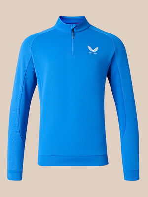 A bright blue long-sleeve quarter-zip athletic pullover with a white wing logo on the left chest, displayed on a solid beige background.