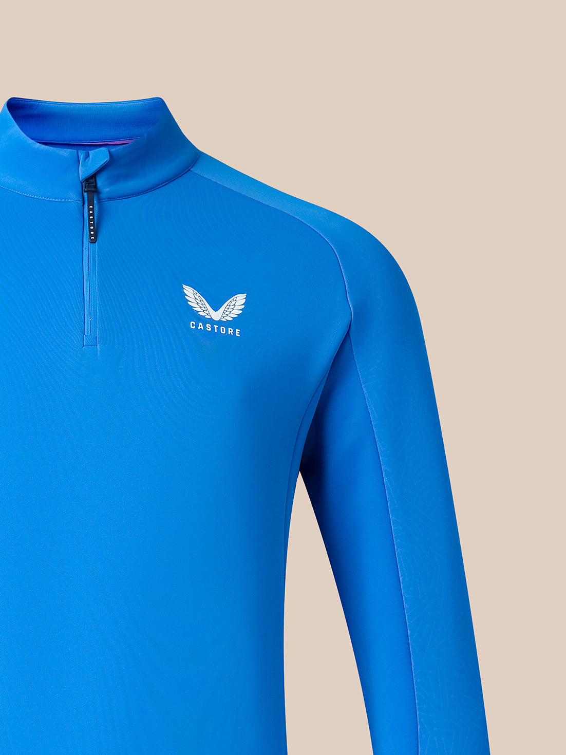 A close-up of a bright blue long-sleeve athletic quarter-zip top with a white winged logo and the word 'CASTORE' on the left chest, shown against a solid beige background.