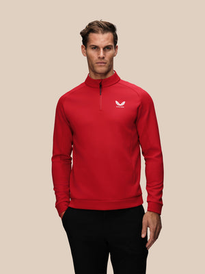 A man wearing a bright red long-sleeve quarter-zip pullover sweatshirt with a small white wing logo on the left chest, paired with black pants, against a solid beige background.