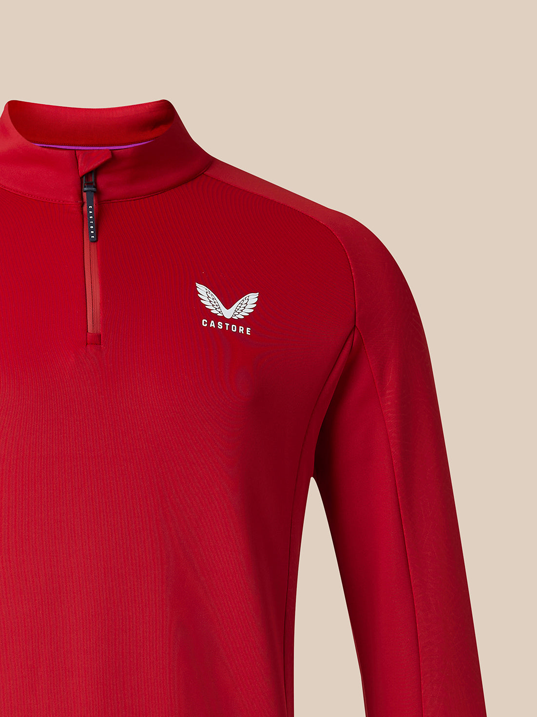 A close-up of a red long-sleeve athletic quarter-zip pullover with a white winged logo and the word 'CASTORE' on the left chest, shown against a solid beige background.