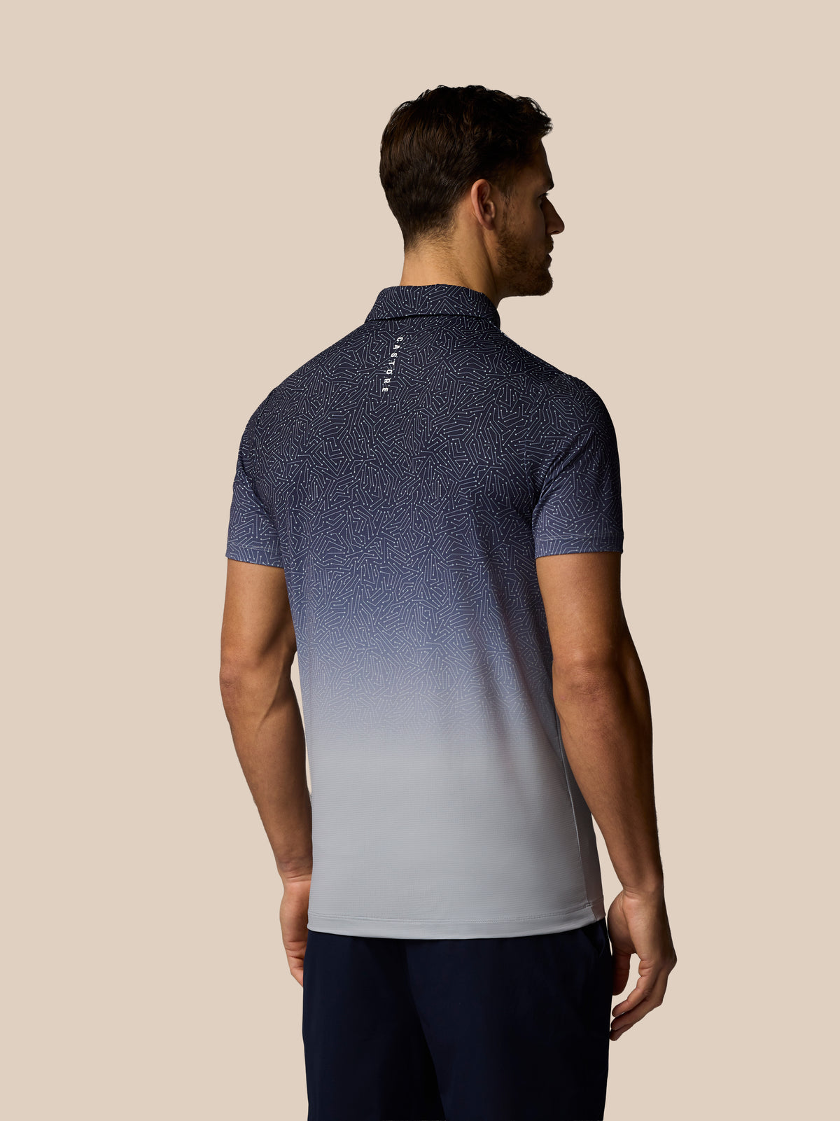 A man shown from the back wearing a short-sleeve polo shirt with a dark blue to light gray gradient and fine speckled pattern, paired with dark navy pants, against a solid beige background.