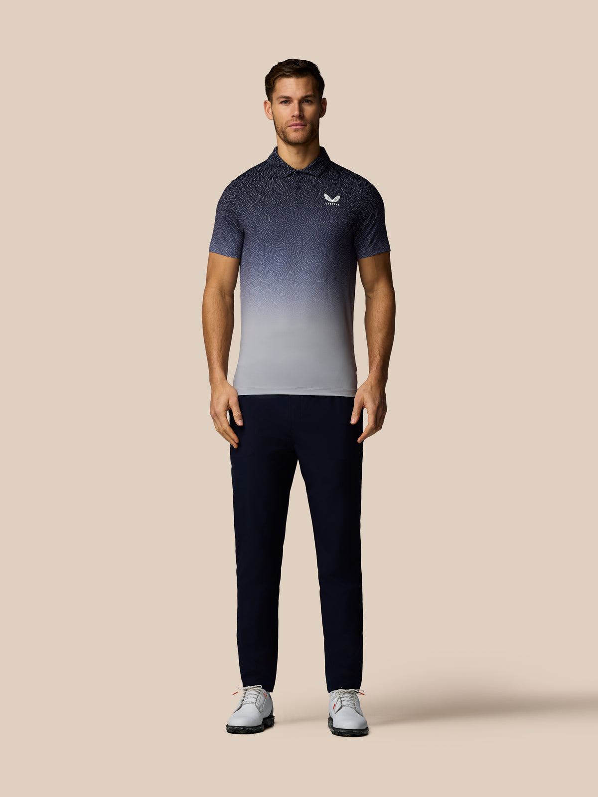 A man modeling a short-sleeve polo shirt with a dark-to-light gradient design, paired with dark trousers and white sneakers, against a solid beige background.