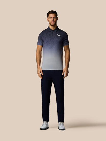 A man modeling a short-sleeve polo shirt with a dark-to-light gradient design, paired with dark trousers and white sneakers, against a solid beige background.