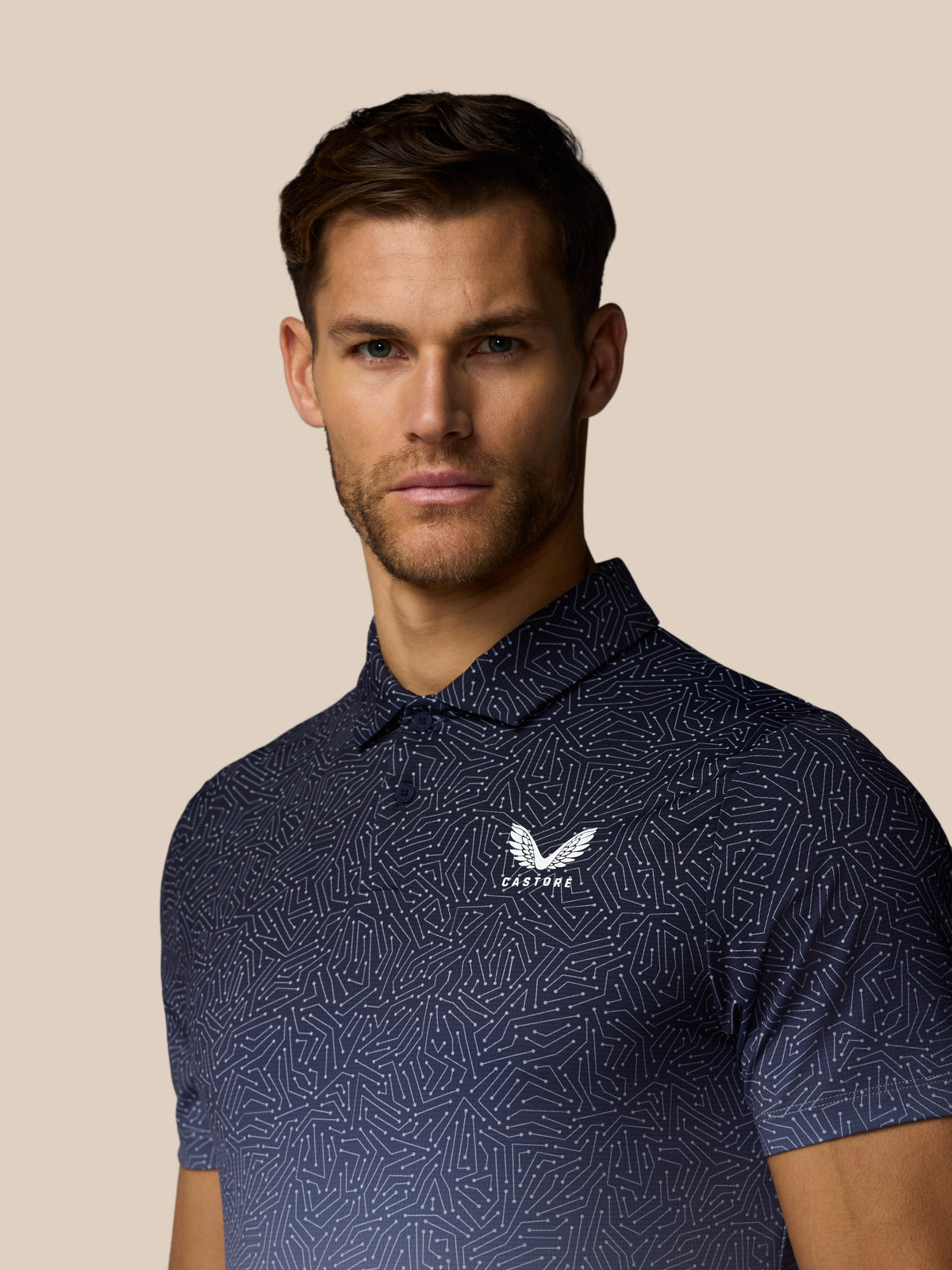 A man wearing a dark navy blue short-sleeve polo shirt with a white geometric pattern and a winged logo on the chest that reads 'Castore'. The background is a solid light beige color.