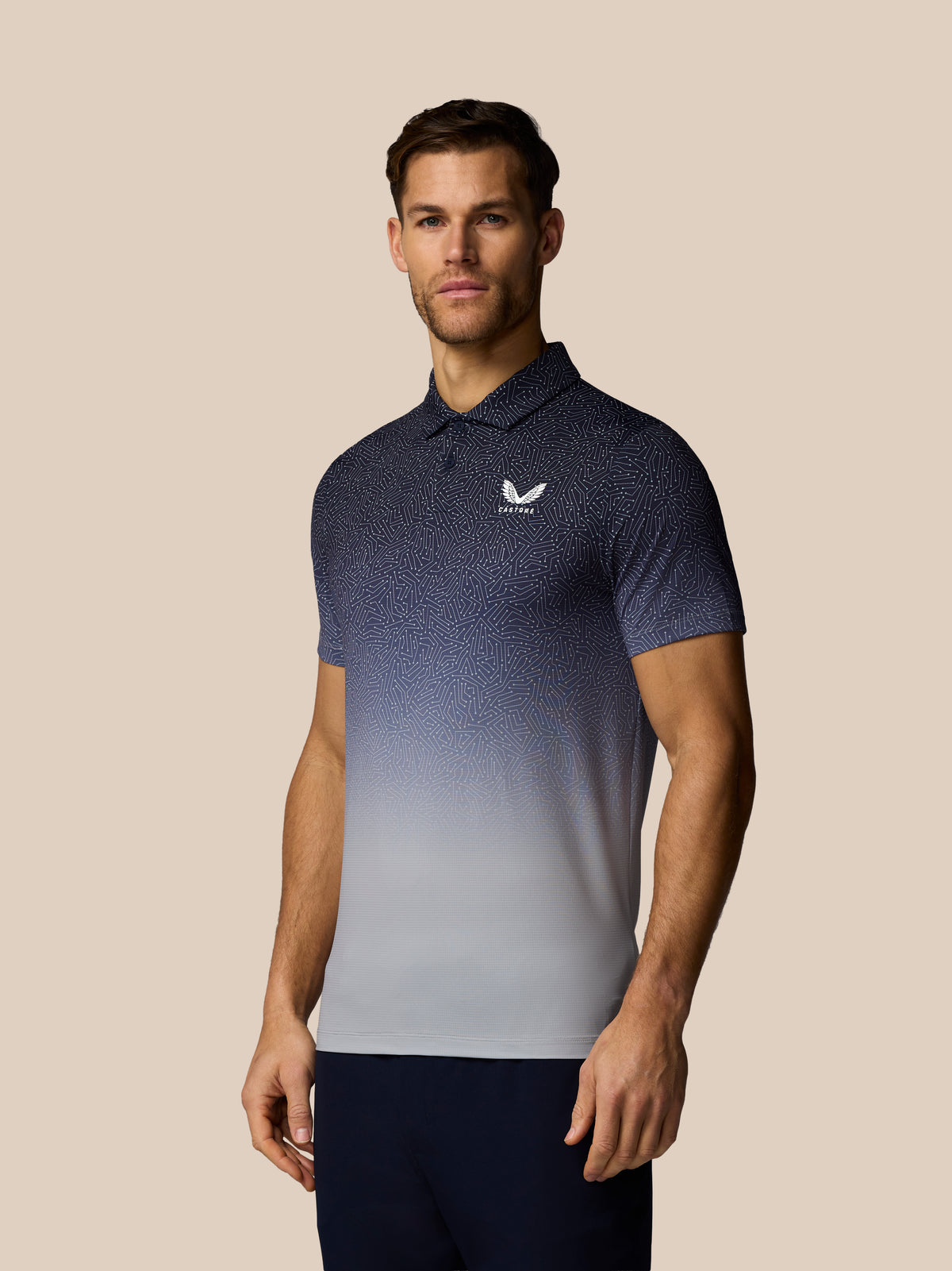 A man wearing a short-sleeve polo shirt with a dark-to-light gradient pattern and a white logo on the chest, standing against a solid beige background.
