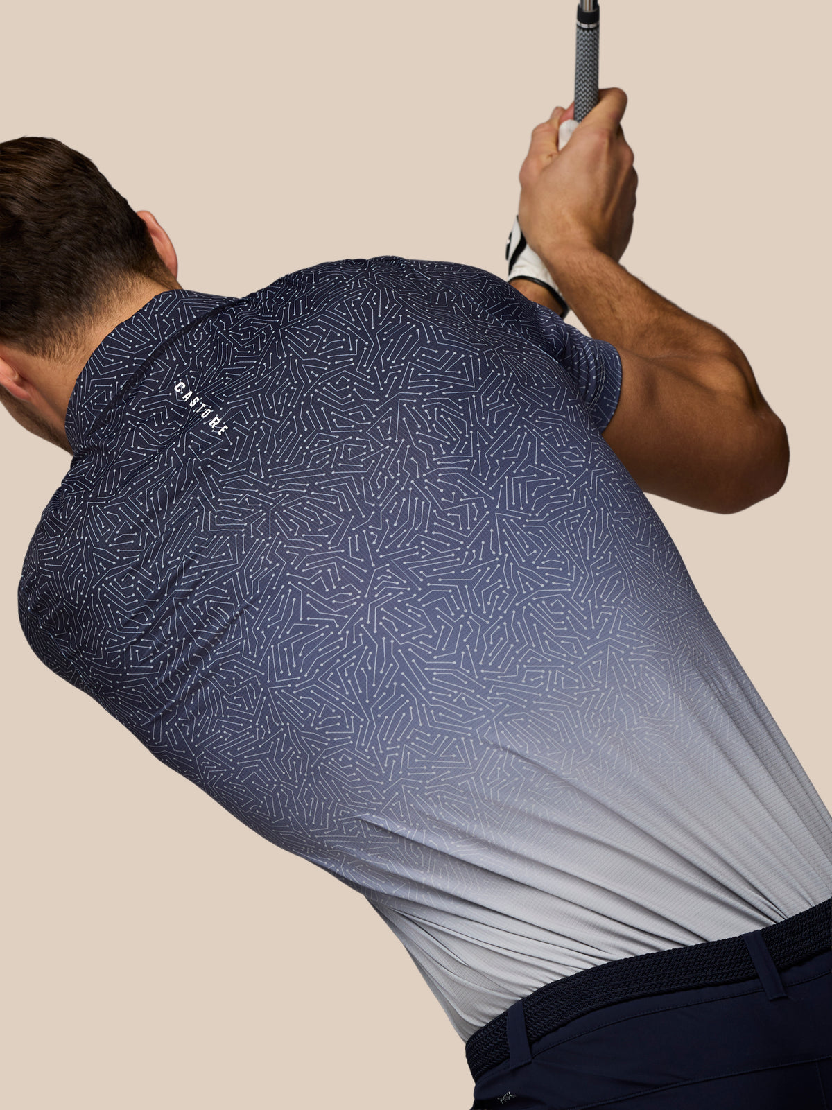 A man wearing a navy blue and gray patterned golf shirt, captured from behind as he swings a club, against a solid beige background.