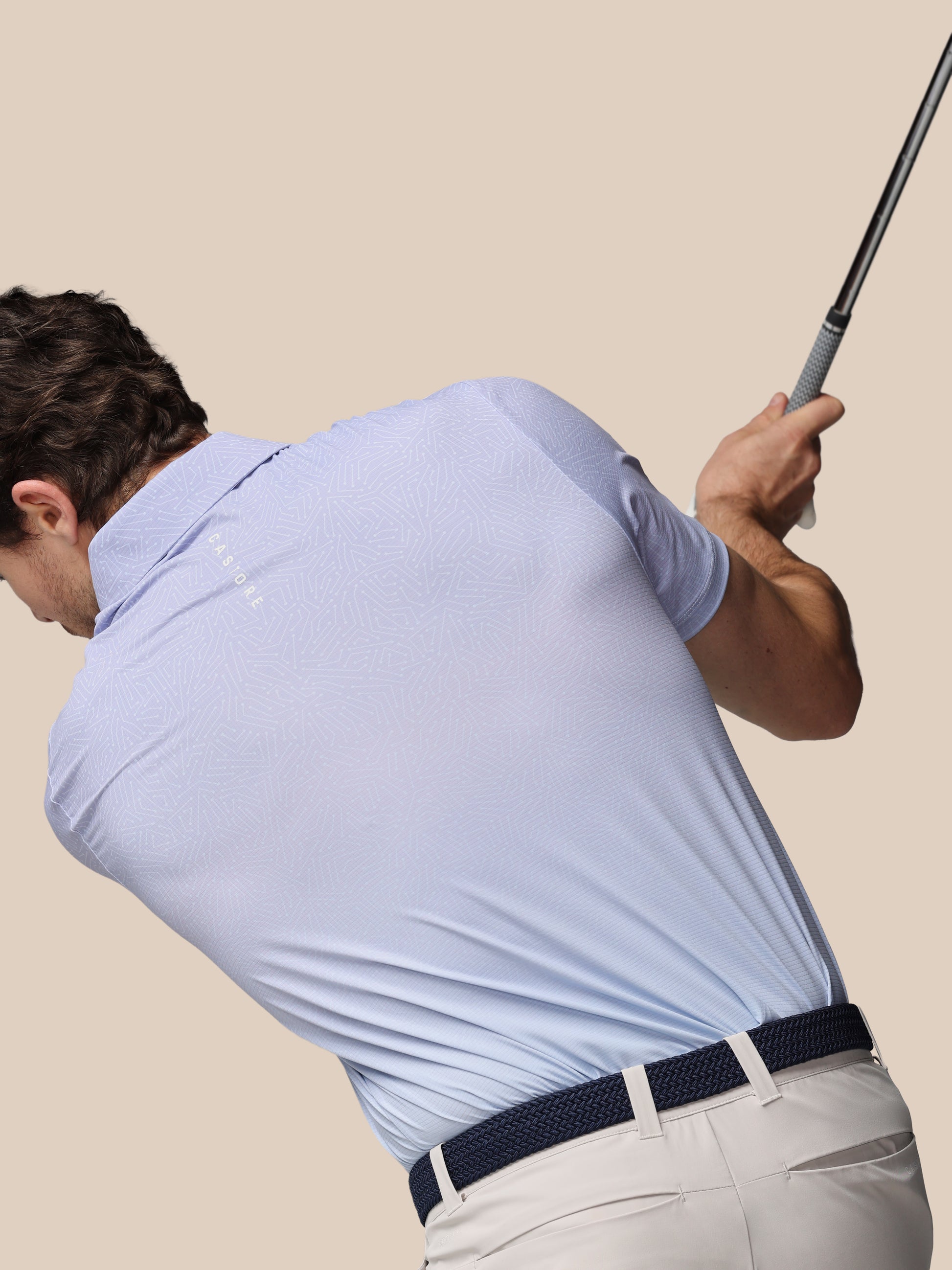Man in light blue shirt and beige pants swinging a golf club against a beige background